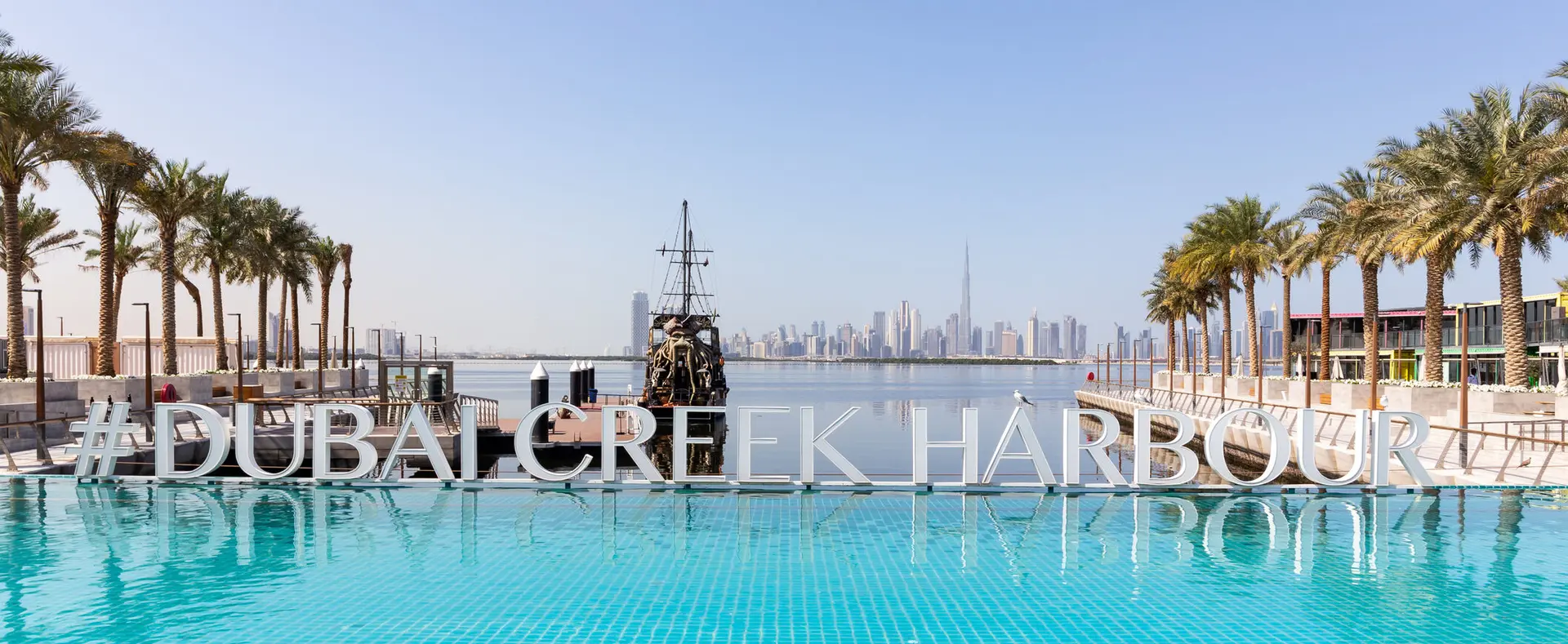 Harbour Promenade waterfront and #DUBAI CREEK HARBOUR sign at Dubai Creek Harbour with Downtown Dubai skyline and Burj Khalifa in the background