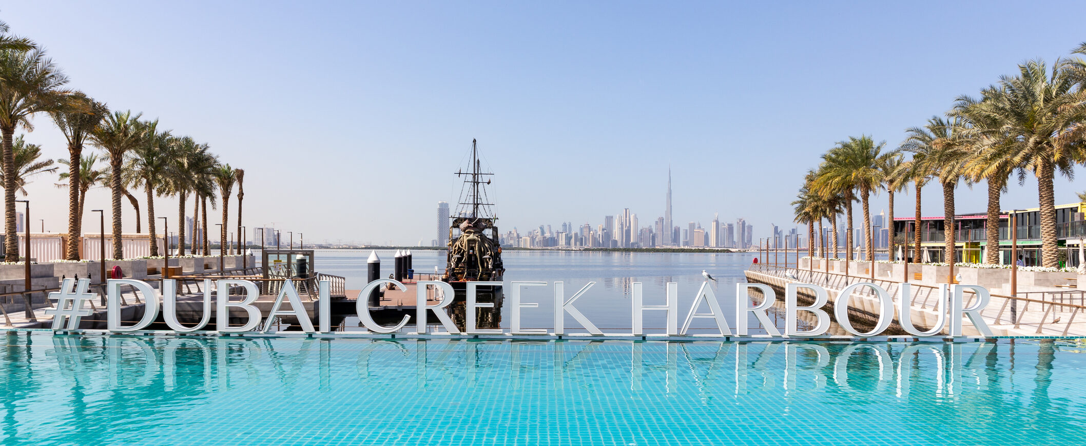 Harbour Promenade waterfront and #DUBAI CREEK HARBOUR sign at Dubai Creek Harbour with Downtown Dubai skyline and Burj Khalifa in the background