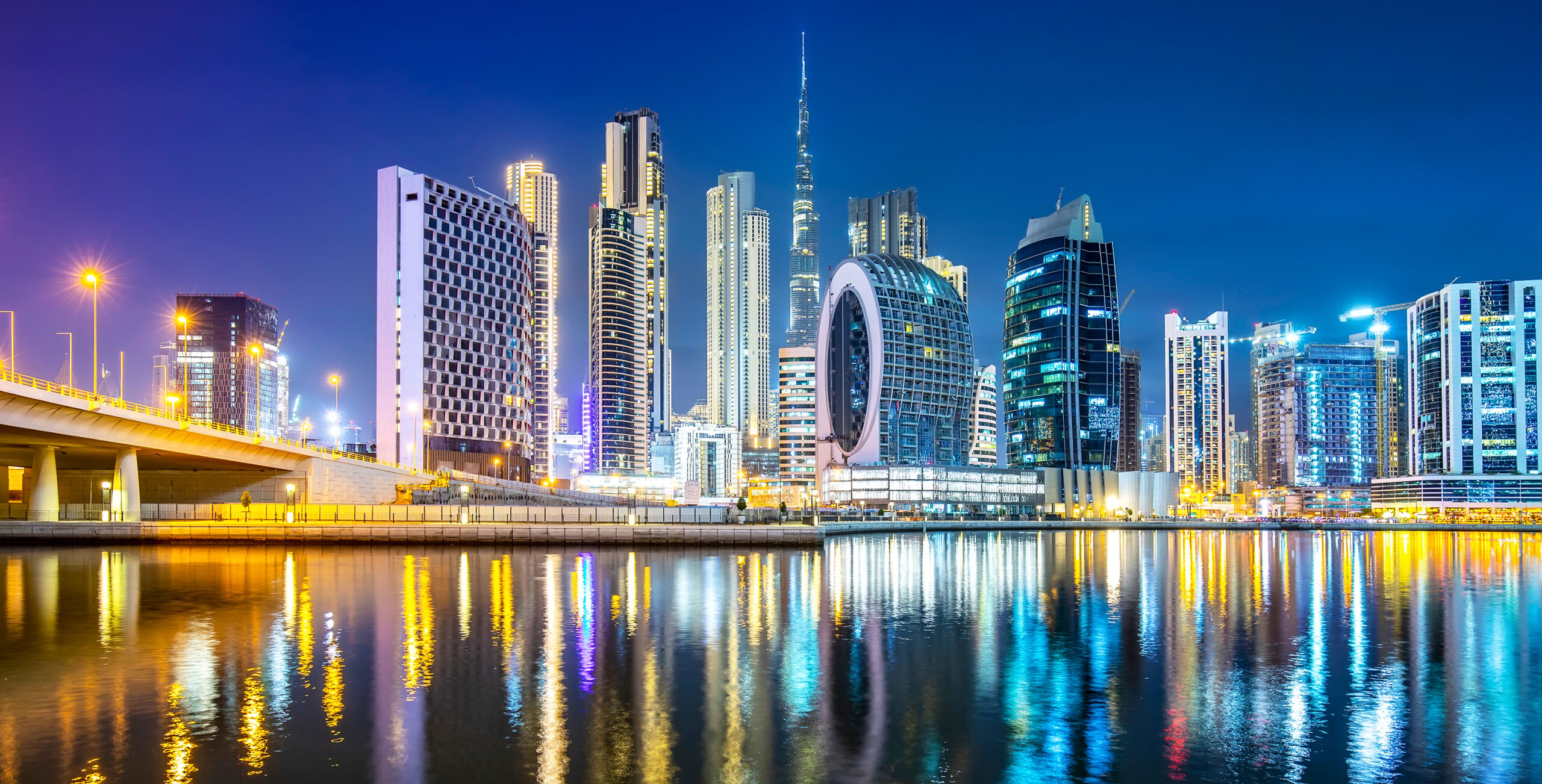 Night skyline of Business Bay, Dubai, with Burj Khalifa and canal reflections.